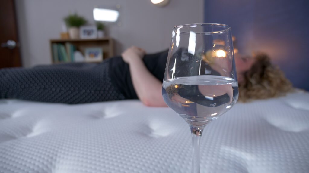 A woman lying on the Plank Firm Luxe mattress next to a wine glass filled with water.