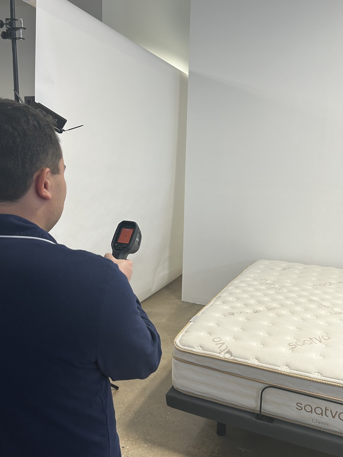 A man standing in front of a Saatva mattress and aiming a thermal gun at its surface to get its temperature.