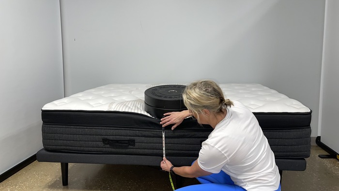 A woman kneeling next to the Nolah Evolution mattress while it has a stack of weights on it. She's measuring the bed's sinkage.
