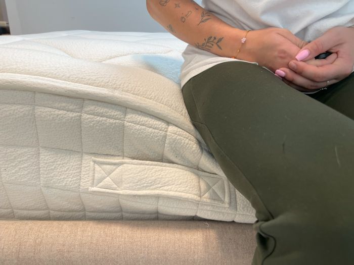 A closeup of the side of the Avocado Luxury Organic mattress. A woman is sitting on it and causing some sinkage to the layers.