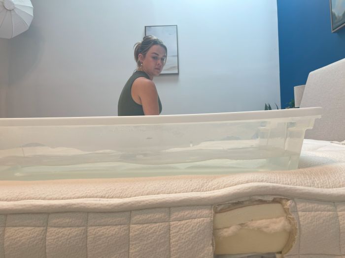 A woman sits on an Avocado Organic mattress next to a bin full of water.