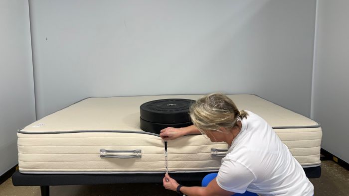 A woman kneeling next to a Nolah Natural mattress. A stack of weight sits on top of the bed and she's measuring the sinkage with a tape measure.
