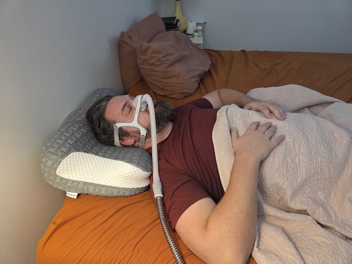 A man sleeping on his back on the Elviros pillow. He's wearing a sleep apnea mask as he dozes.