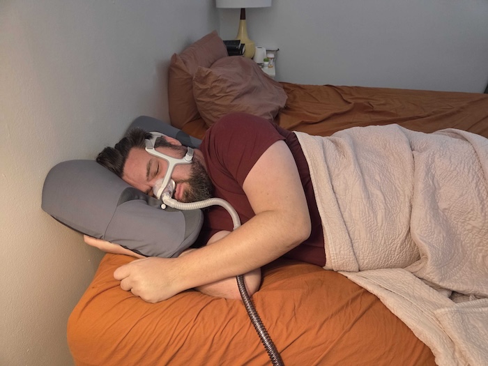 A man sleeping on his side on the Ikstar pillow. He's wearing a sleep apnea mask as he dozes.