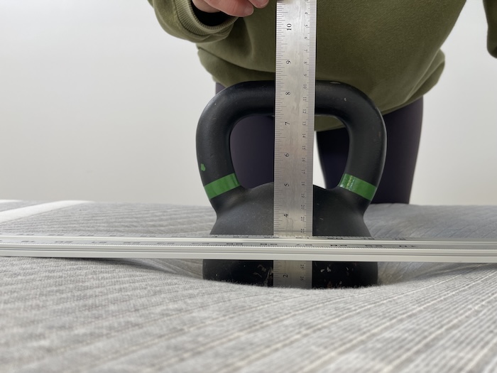 A kettlebell resting on a Leesa mattress. A woman is using a ruler to measure the sinkage.