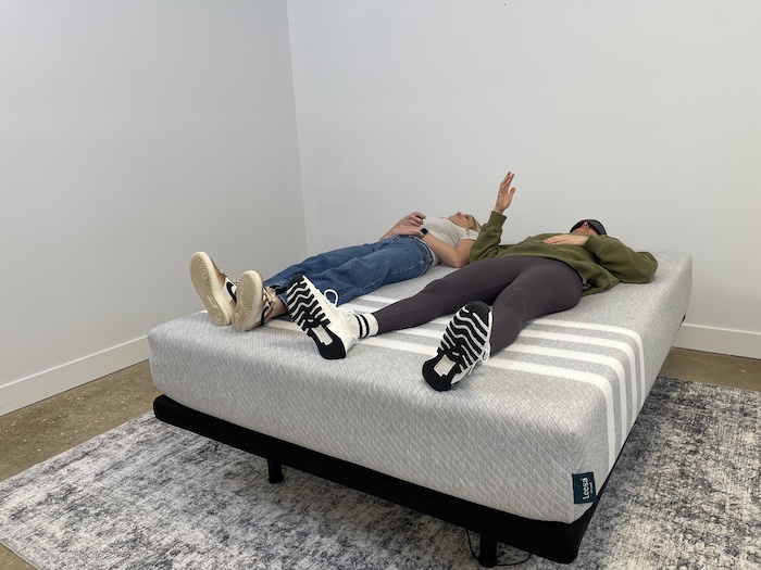 Two women lying next to each other on the Leesa Original mattress. One of them is wearing a blindfold and using their hand to signal how much they can feel the other one's movement.