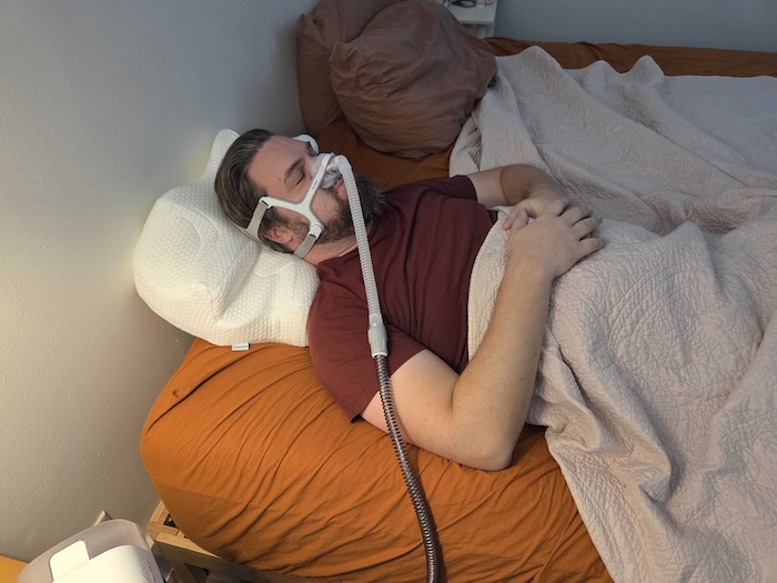 A man sleeping on his back on the Lunderg pillow. He's wearing a sleep apnea mask as he dozes.