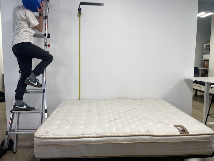 A many climbing a ladder next to the Saatva mattress with a medicine ball in his hand. He's about to drop it on the Saatva to test the bed's bounce.