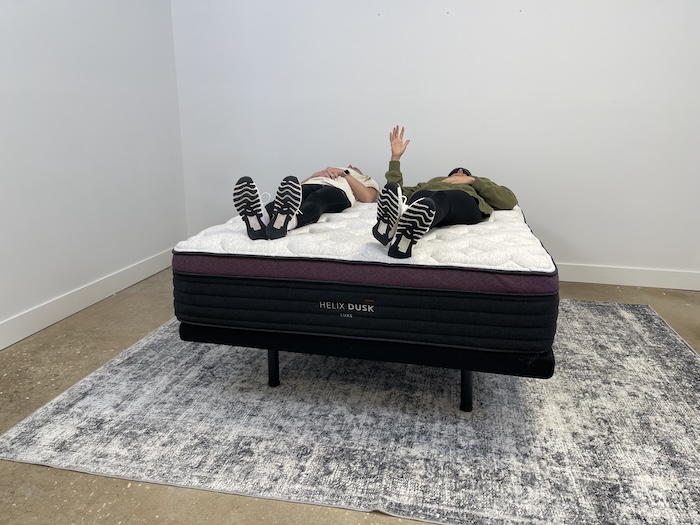 Two women lying side by side on the Helix Dusk Luxe mattress. One of them is wearing a sleep mask and raising her hand to indicate that she can feel the movement of her partner.